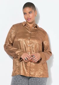 Bronzefarbene, metallische Button-Up-Bluse mit einer Brusttasche, langen Ärmeln und einem lockeren Schnitt. Kombiniert mit grauen Leopardmusterhosen, die Ringe und orangefarbene Nägel haben.