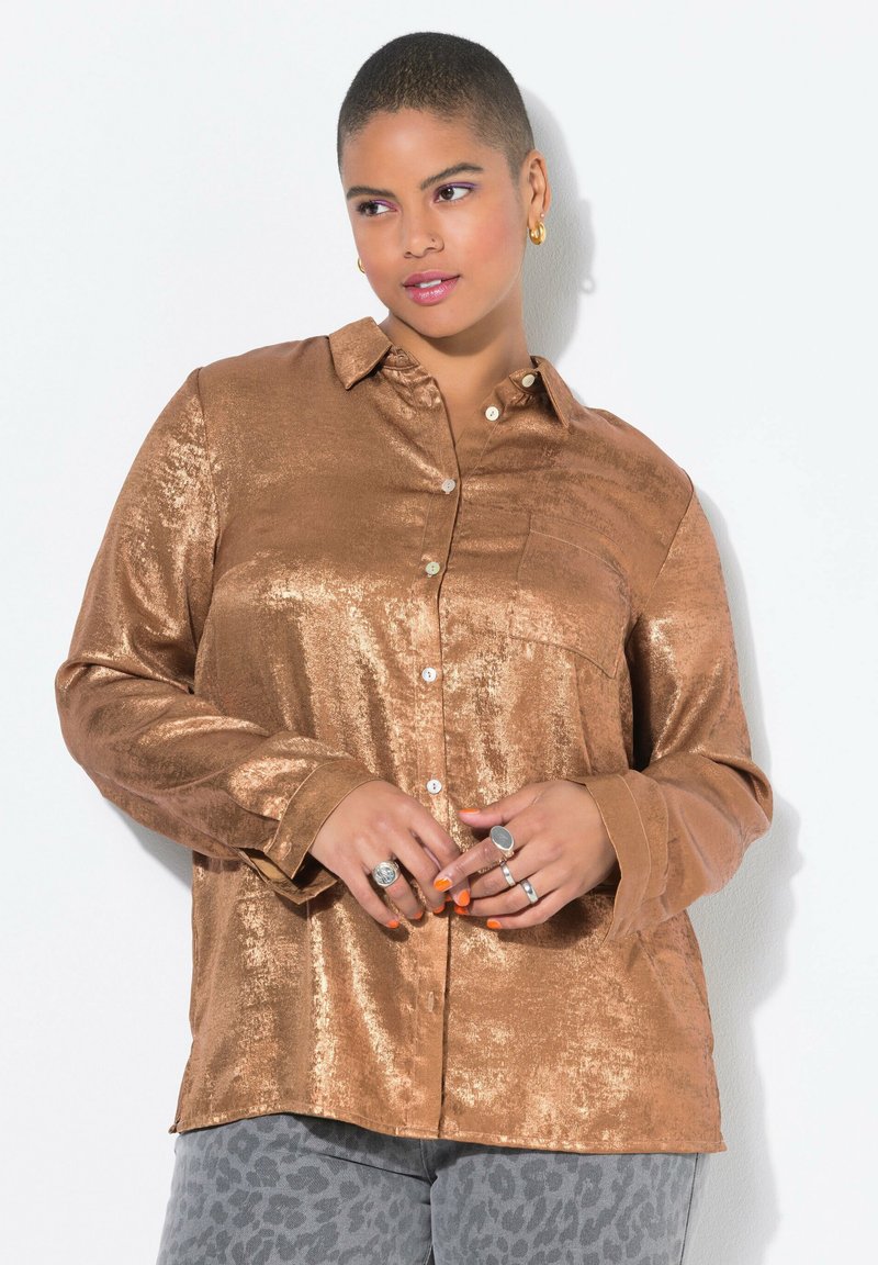 Bronzefarbene, metallische Button-Up-Bluse mit einer Brusttasche, langen Ärmeln und einem lockeren Schnitt. Kombiniert mit grauen Leopardmusterhosen, die Ringe und orangefarbene Nägel haben.
