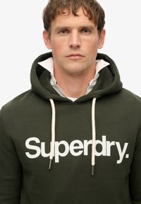 Felpa verde con logo "Superdry." bianco, cordini e una finitura texturizzata. Indossata sopra una camicia bianca con colletto e polsini in vista.