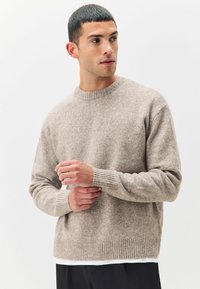 Suéter de punto beige con cuello redondo, puños y dobladillo acanalados, y un acabado de textura suave. Capa sobre una camisa blanca.
