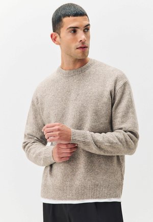Beige gebreide trui met een ronde hals, geribde manchetten en boord, en een zachte textuur. Gelaagd over een wit shirt.