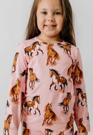 Smiling young girl wearing pink pajamas with brown and white horses running printed all over the fabric against a plain background.