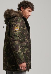 Hombre con chaqueta de camuflaje verde y marrón con capucha forrada de piel, de pie de perfil contra un fondo gris claro.