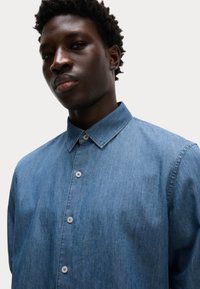 Camicia in denim di colore azzurro chiaro, con colletto button-down e chiusura con bottoni sul davanti. Il tessuto appare morbido con una superficie liscia.