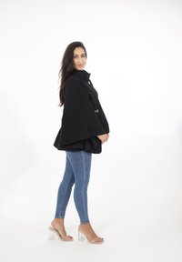 Svart cape-liknande kappa med hög krage och silverdetaljer, i kombination med blå skinny jeans och klara blockklack-sandaler på en vit bakgrund.