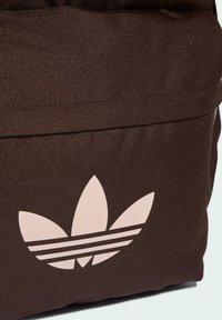 Brązowa torba z tkaniny o gładkiej fakturze i wyraźnym, jasnoróżowym logo koniczyny, z trzema poziomymi paskami pod logo.