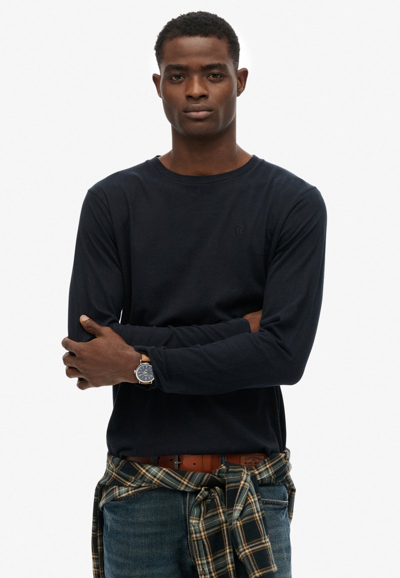 Navyblauwe longsleeve shirt gemaakt van zachte stof, gecombineerd met een blauwe spijkerbroek. Een geruit flanellen shirt is om de taille geknoopt. Heeft een polshorloge.