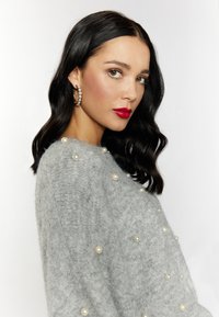 Pulover gri din tricot, decorat cu mici perle albe, având o croială lejeră și mâneci largi. Modelul poartă cercei în cerc și ruj roșu.