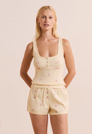 Femme blonde portant un débardeur côtelé jaune pâle et un short assorti avec de petits imprimés floraux roses, debout devant un fond beige.
