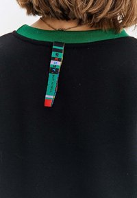 Černá mikina s zeleným límcem a barevnou nášivkou vzadu s textem "NEPOŘÁDNÝ ALE ŠŤASTNÝ." Měkká látka, volný design.