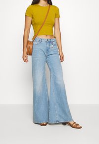 Geel cropped t-shirt, lichtblauwe flared jeans en een bruine schoudertas. Sandalen met meerdere banden. Simpel ontwerp, casual pasvorm.