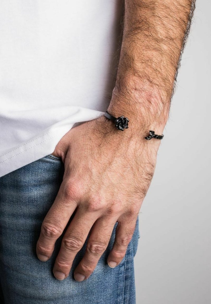 Main gauche d'un homme reposant dans la poche d'un jean bleu, portant un bracelet noir avec un détail en forme de rose au poignet, chemise blanche visible.