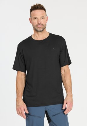 Man met kort haar en baard, gekleed in een zwart T-shirt en blauwe broek, staand tegen een effen witte achtergrond.