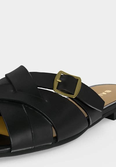 Sandale plate en cuir noir avec larges brides croisées et boucle carrée dorée sur une semelle intérieure beige.