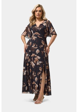 Kvinde i en lang sort blomsterkjole med flagrende ærmer og slids højt oppe på låret, iført guld høje hæle sandaler og et armbånd.