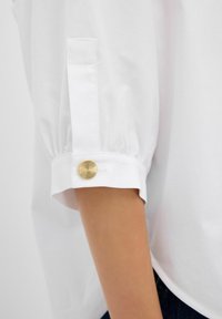 Hvid bluse med puffede ærmer, der har en guld dekorativ knap ved mansjetten og en glat stofstruktur. Casual pasform og enkelt design.