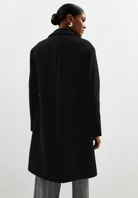 Zwarte wollen jas met een op maat gemaakte silhoutte, grote kraag en lange mouwen; gladde textuur met een rechte snit die onder de heupen valt.