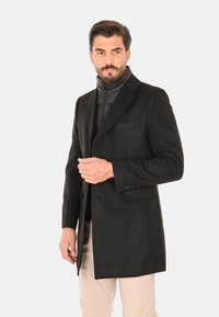 Uomo con barba che indossa un cappotto nero sopra una giacca nera con zip e pantaloni beige, in piedi mentre si sistema il cappotto con una mano.