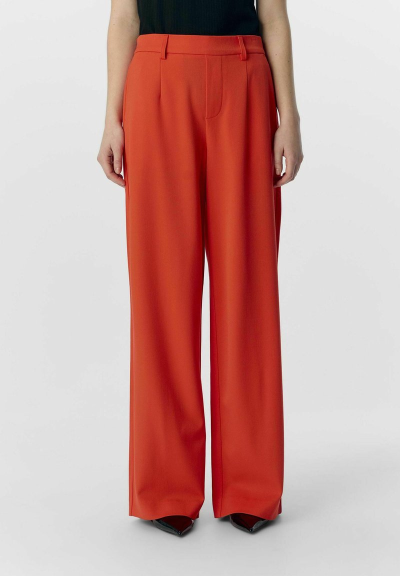 Pantalons rouges à jambes larges en tissu lisse avec taille haute, dotés de poches avant et d'un look ajusté. Associés à des chaussures noires à bout pointu.