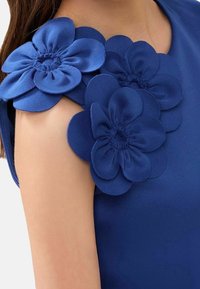 Sininen mekko, jossa on kolme suurta, kerrostettua samettikukkasta olkapäällä. Saumaton pinta, vartalonmyötäinen muotoilu ja ei näkyviä kiinnityksiä.