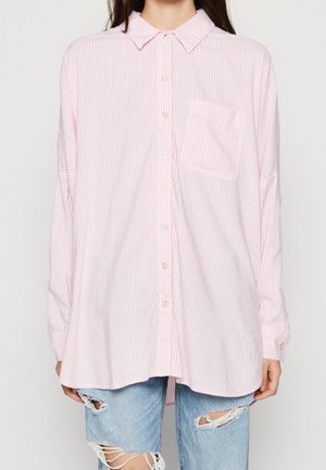 Długa koszula na guziki w jasnoróżowe i białe paski z kieszenią na piersi, noszona bez włożenia w spodnie, na tle podartych jasnoniebieskich jeansów.