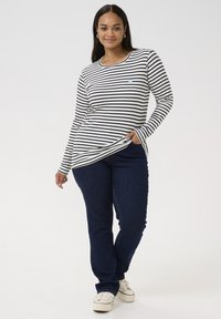 Chemise à manches longues rayée noir et blanc avec un col rond, assortie à un jean bleu foncé et des baskets blanches. Logo subtil sur la poitrine.