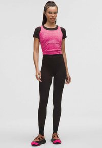 Top sportivo aderente di colore rosa brillante con tessuto texturizzato, abbinato a leggings neri a vita alta e scarpe da ginnastica multicolori.