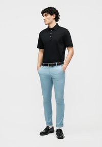 Jeune homme debout, les mains dans les poches, portant un polo noir, un pantalon slim bleu clair, une ceinture noire, des chaussettes blanches et des mocassins noirs.