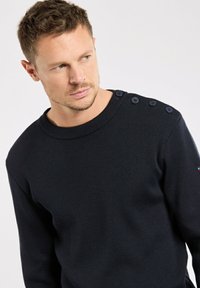 Mann mit kurzen Haaren, der einen schwarzen Strickpullover mit vier Knöpfen an der linken Schulter trägt und leicht nach rechts schaut.