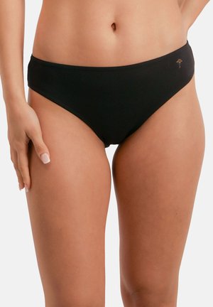 Nainen, jolla on yksinkertaiset mustat bikinitrikoiset ja pieni kultainen merkki oikeassa lonkassa, seisoo valkoista taustaa vasten.