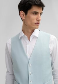 Hellblaues Vest aus strukturiertem Stoff, mit V-Ausschnitt, drei Knöpfen und zwei Seitentaschen, getragen über einem weißen Hemd.