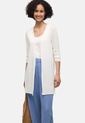 Kvinde med krøllet hår iført hvid cardigan med lange ærmer, hvid top, blå løstsiddende bukser, smilende, holder en brun håndtaske.