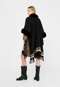 Donna che indossa un cappotto nero con frange e rifiniture in pelliccia sopra un vestito marrone e stivaletti neri alla caviglia, in piedi su uno sfondo bianco semplice.