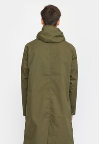 Olivengrüne, lange Jacke mit kapuze, verfügt über eine glatte Außenseite, geraden Schnitt und eine Naht in der Mitte. Minimalistisches Design, keine sichtbare Hardware.