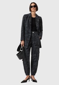 Blazer e pantaloni con motivo camouflage in tonalità grigie scure, abbinati a una maglietta nera e eleganti scarpe con tacco nere, mentre tiene una piccola borsa a tracolla.