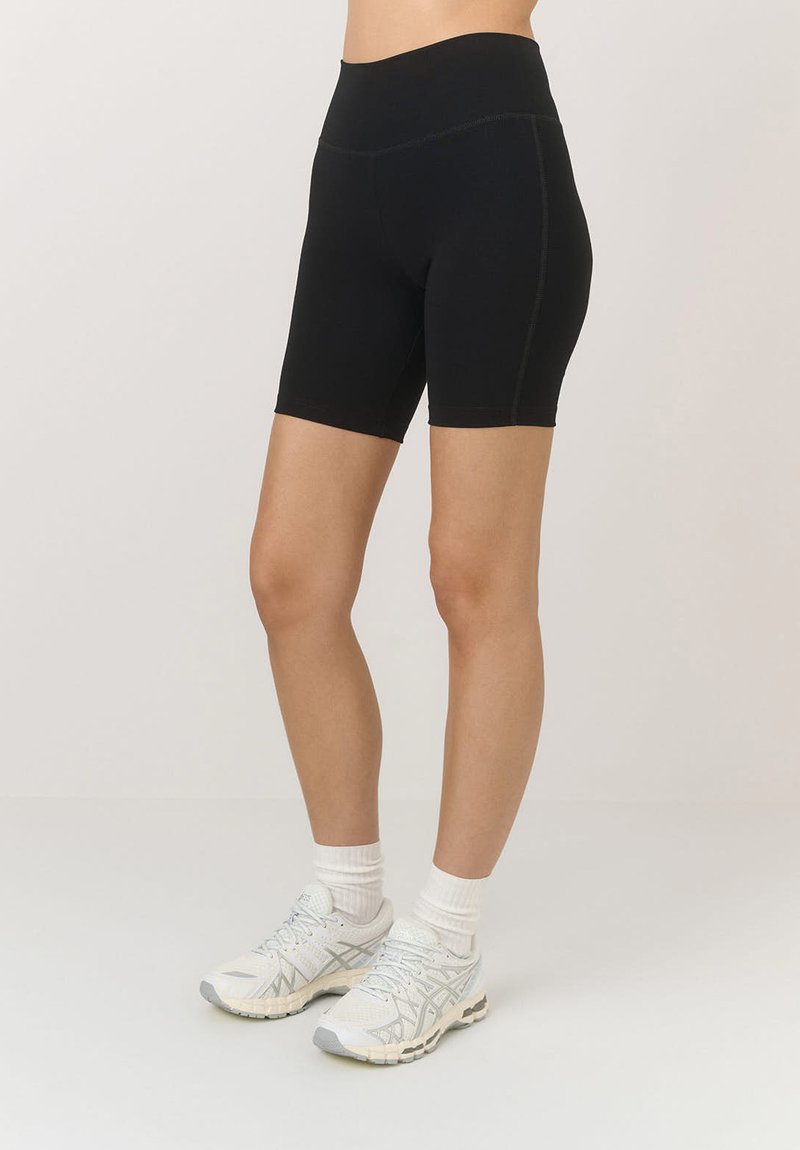 Czarne rowerowe szorty z wysokim stanem wykonane z elastycznego materiału, z płaskimi szwami i długością do połowy uda, w połączeniu z białym obuwiem sportowym.