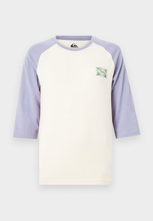 Krém színű ing, levendula színű háromnegyedes raglán ujjakkal és egy kis zöld-fehér napsugaras logóval a mellkasán.