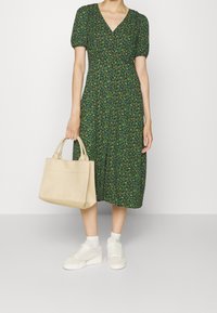 Femme portant une robe midi à fleurs vertes, tenant un sac fourre-tout beige, associée à des chaussettes blanches et des baskets blanches, debout devant un mur uni.