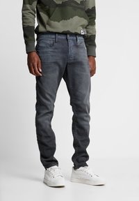 Slim-fit jeans i mörkgrå denim med en subtil blekt textur, fem fickor och en enkel knappstängning, paired med vita sneakers.