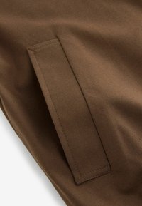 Brauner Stoff mit einer glatten Textur und feiner Rippung. Verfügt über eine rechteckige Tasche mit sauberer Naht, die in das Design integriert ist.
