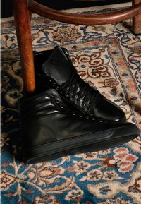 Czarne, skórzane sneakersy za kostkę z teksturowaną boczną panelem, metalowymi oczkami, sznurowadłami i gumową podeszwą, ustawione w pobliżu drewnianego krzesła.