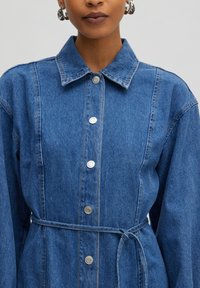 Jeans jurk met een kraag, lange mouwen, zilveren knopen en een bijpassende riem in de taille. Blauw stof met stikseldetails.