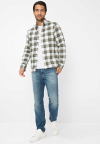Camicia di flanella con motivo a quadri verde, bianco e nero, con bottoni sul davanti, abbinata a jeans blu e sneakers bianche su uno sfondo neutro.