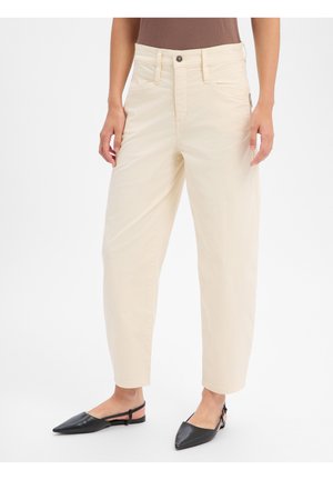 Femme portant un pantalon crème taille haute et raccourci avec poches avant et des ballerines noires à bout pointu avec bride arrière, debout devant un fond blanc.