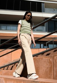 Grünes und weiß gestreiftes ärmelloses Oberteil, beige weite Hosen und weiße Plateausandalen; Figur, die braune Steinstufen hinuntergeht.