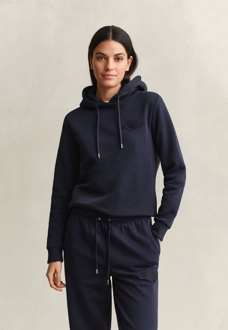 Vrouw die een donkerblauwe hoodie en bijpassende joggingbroek draagt, staand met één hand in de zak tegen een effen lichte achtergrond.