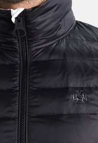 Svart vadderad jacka med hög krage, framdragkedja och subtil broderad logotyp. Materialet verkar slätt med en lätt lyster.