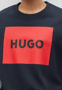 Sudadera de cuello redondo en color navy que presenta un gran rectángulo rojo con la palabra "HUGO" en letras negras y en negrita. Tela suave con un corte holgado.