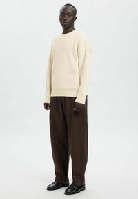 Kreemikas soonikkoes kampsun langevate õlgadega, kombineeritud lahtiste pruunide pükstega ja mustade jalanõudega, neutraalse taustaga.