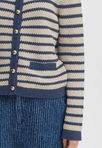 Pruhovaný cardigan v krémové a námořnické barvě, pletená textura s žebrovanými manžetami, pěti zlatými knoflíky a malou přední kapsou. Spojený s modrými pruhovanými kalhotami.